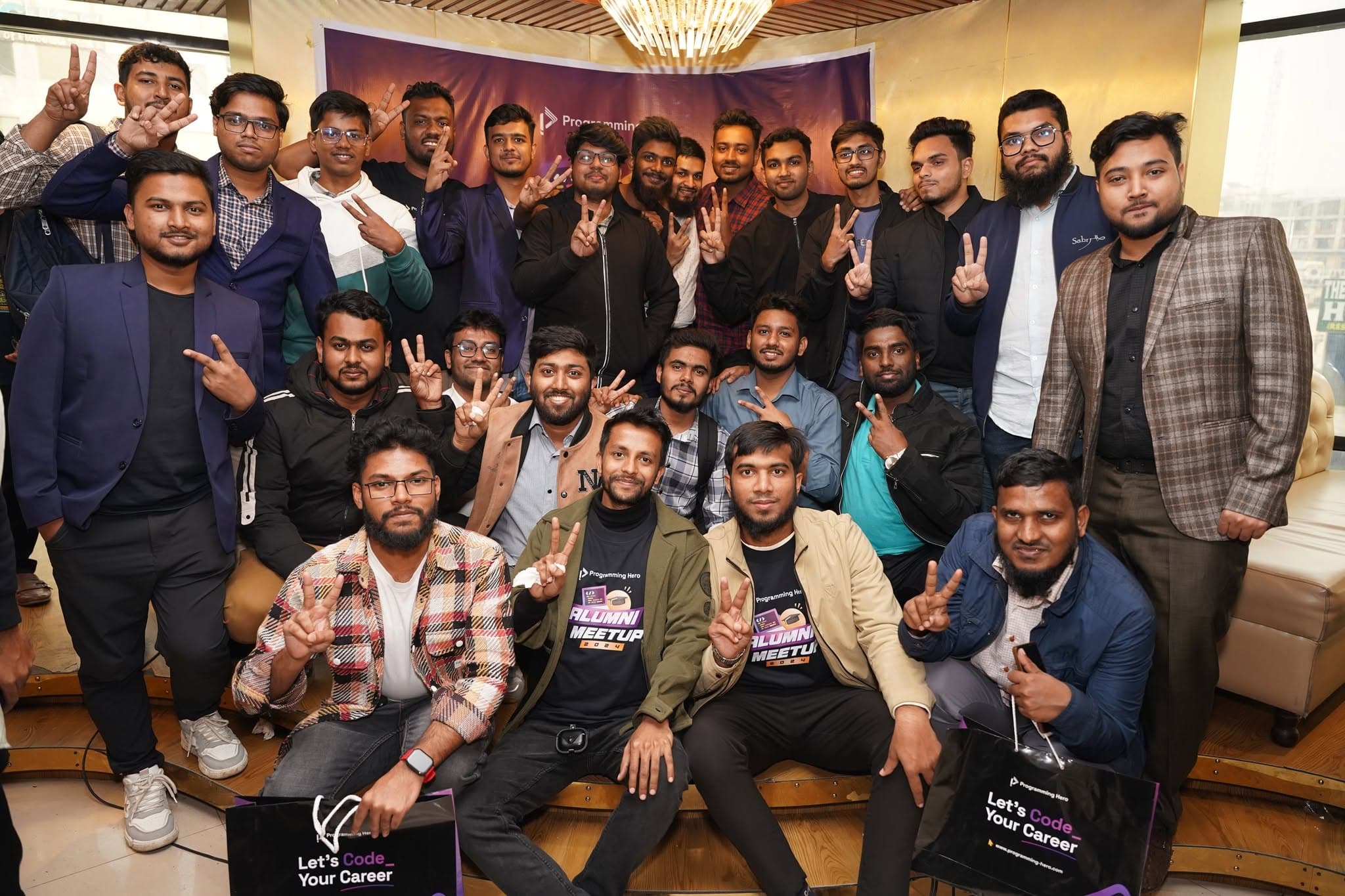 About thirty developers posed together in front of a Programming Hero banner at the 2024 alumni meetup, several wearing Alumni Meetup 2024 shirts and holding Let’s Code Your Career tote bags.