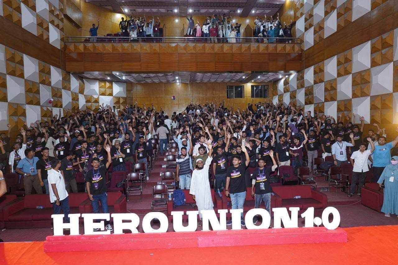 A packed auditorium at HeroUnion 1.0 with several hundred attendees standing with their arms raised, large HEROUNION 1.0 letters on the stage floor in the foreground.